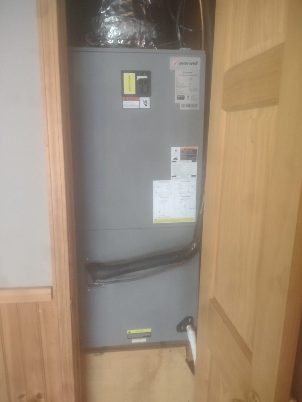 HVAC system installed in a utility closet, with visible ductwork and labels on the front panel, partially obstructed by an open wooden door.