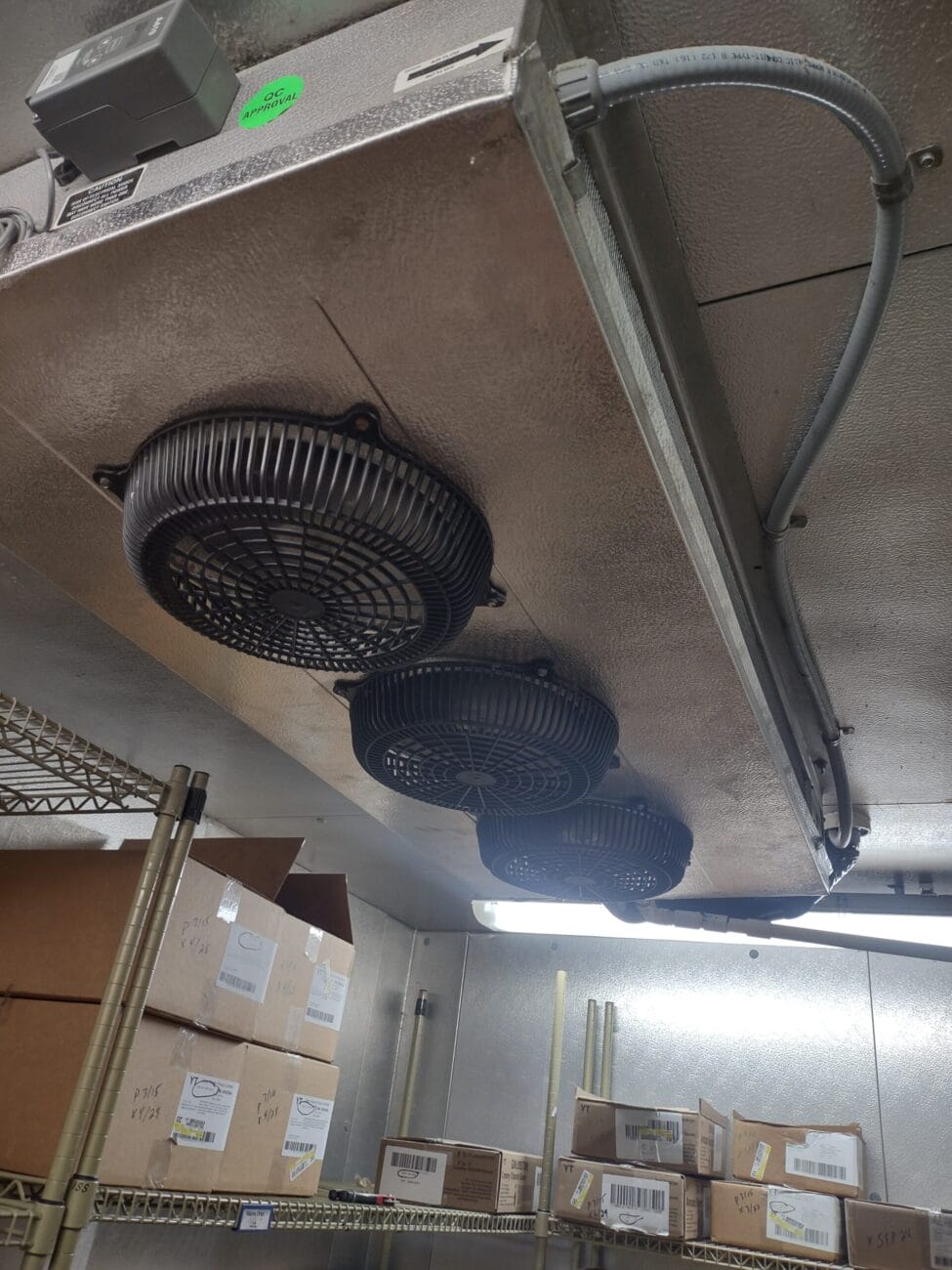 A metal ceiling with three black industrial fans in a storage room. Shelves are loaded with cardboard boxes and items.